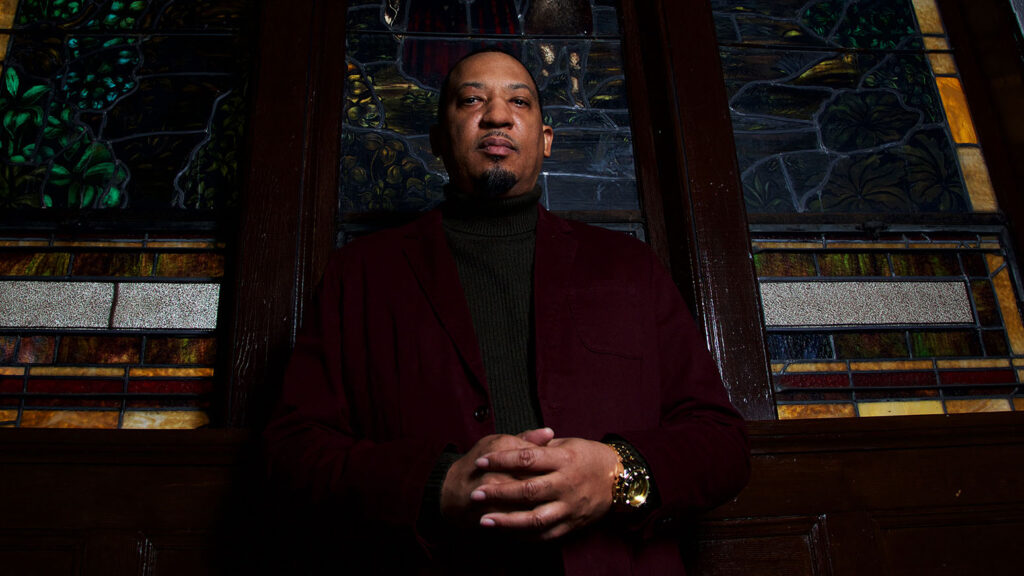 Dasan Ahanu, keynote speaker for Symposium on Afrofuturism and Diasporic Scholarship, stands in front of a stained glass window with his hands folded