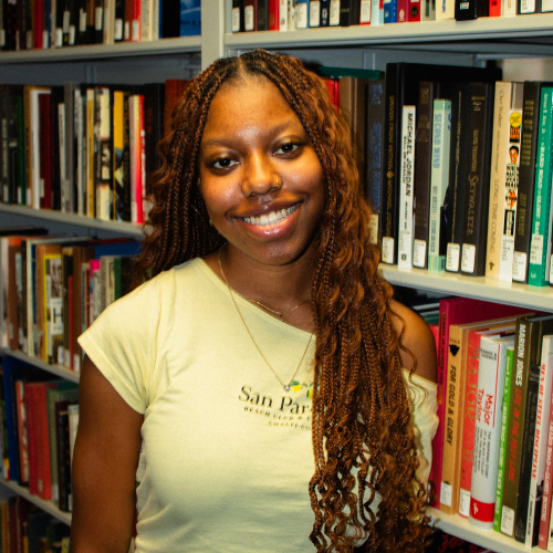 Young woman with long braided hair smiles warmly in a library, wearing a light-colored shirt. Bookshelves filled with various books surround her.
