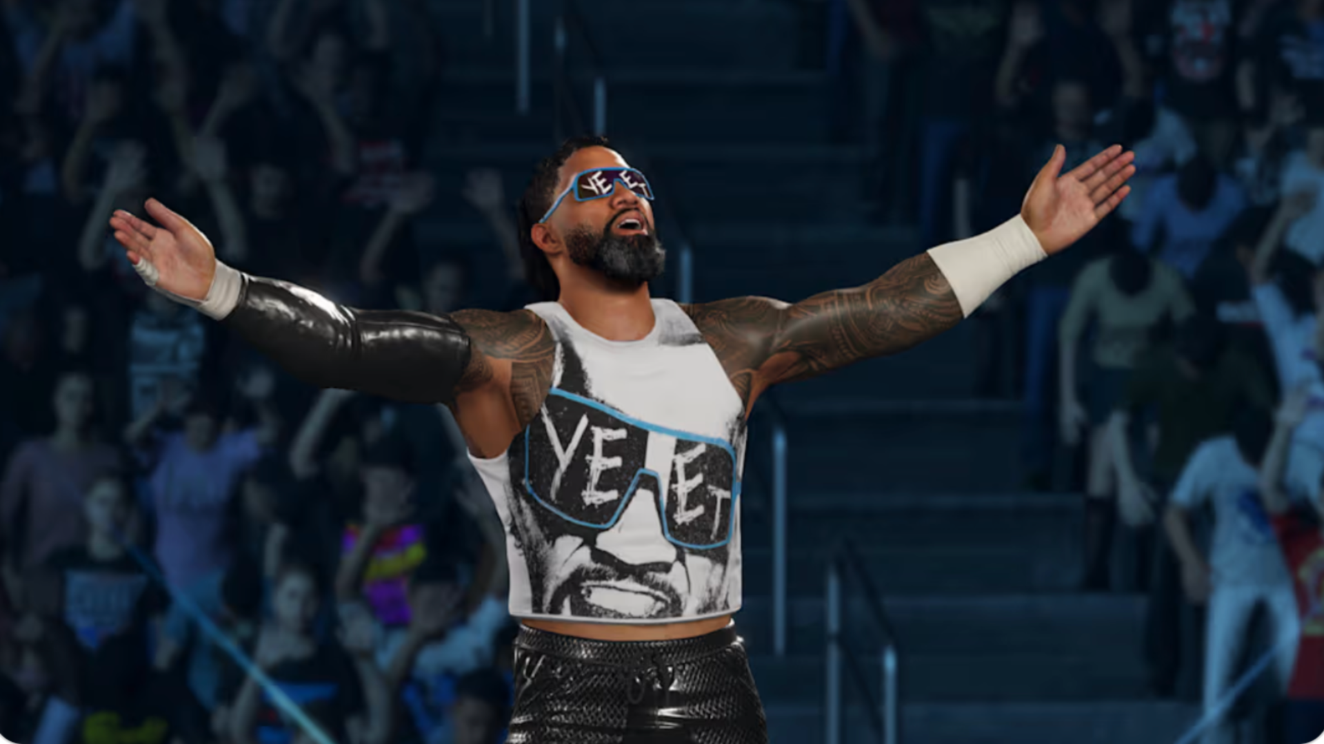 A wrestler with tattooed arms and sunglasses poses confidently with arms outstretched, wearing a graphic tank top in front of a cheering crowd.